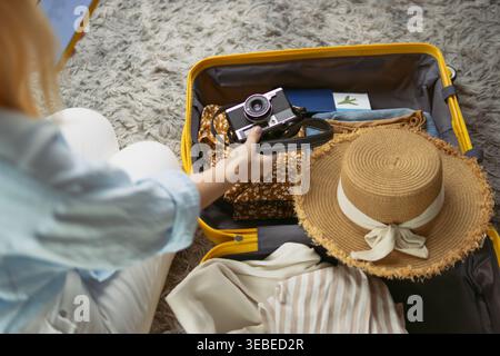 Frau, die einen gelben Koffer mit Sommerkleidung, gestreiftes Hemd, Strohhut und Retro-Kamera neben Pass und Tablet auf einem flauschigen grauen Teppich packt, Top VI Stockfoto