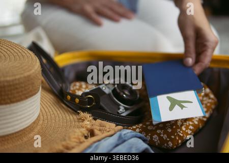 Die Hand der Person legt einen Reisepass mit einem Flugzeugsymbol in einen offenen Koffer, verpackt mit einer Vintage-Kamera, einem Strohhut und Kleidung, bereit für die Fahrt Stockfoto