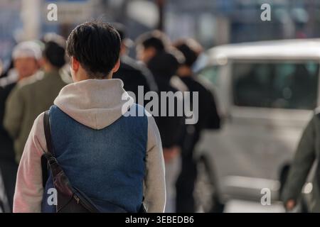 Junger Mann in mehrschichtiger Streetwear aus Jeansweste und Hoodie, der in der Menge auf der belebten Stadtstraße spaziert. Die offene Szene fängt Anonymität und den Alltag ein. Stockfoto