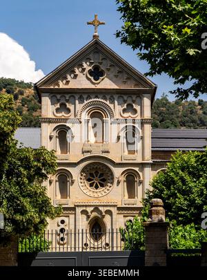 Vorderansicht einer kunstvollen historischen Kirchenfassade mit Kreuz, Bogenfenstern und Rosenfenster, eingerahmt von grünen Bäumen unter blauem Himmel. Stockfoto