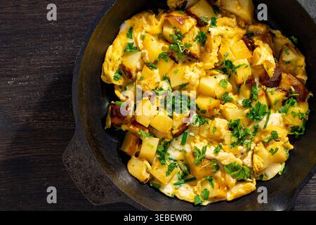 Gebratene Kartoffeln mit Eiern und Petersilie in der Pfanne. Hausgemachte Kartoffelkartoffeln mit Rührei und frischen Kräutern in schwarzer Pfanne auf dunklem Holz. Rustikaler Urlaub Stockfoto