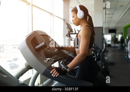 Junge Schwarze Frau passt Laufbandeinstellungen im Fitnessstudio an und trägt weiße Kopfhörer Stockfoto