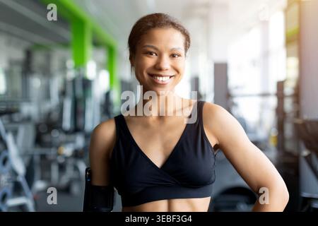 Lächelnde junge Schwarze Frau in schwarzem Sport-BH im hellen Fitnessstudio Stockfoto
