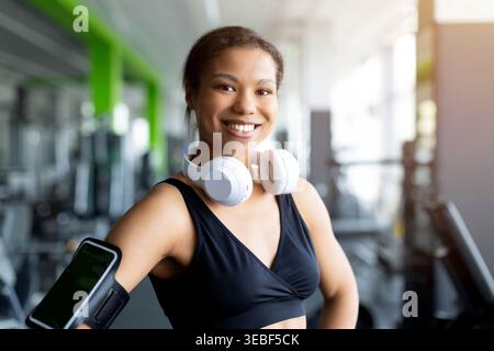 Fröhliche junge schwarze Frau, die weiße kabellose Kopfhörer trägt und im Fitnessstudio lächelt Stockfoto