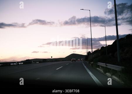 Autos fahren entlang einer malerischen Küstenstraße, während die Sonne untergeht, werfen einen sanften Glanz auf den Himmel und beleuchten den Weg vor Ihnen mit Straßenlaternen Stockfoto