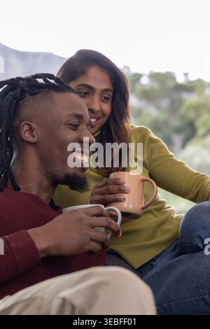 Afroamerikaner und Indianerin sitzen auf dem Sofa und tragen Pullover, die Keramikbecher lächelnd halten Stockfoto