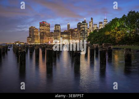 Bildunterschrift: Alte Holzpfeiler im East River mit der beleuchteten unteren Skyline von New York City während der blauen Abenddämmerung Stockfoto