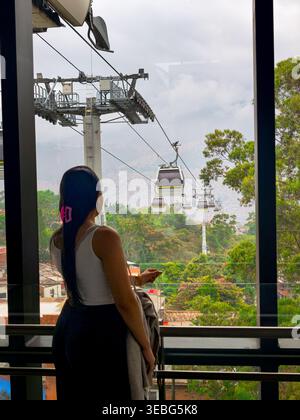 Rückansicht einer Frau, die an einem Fenster steht und Seilbahnen ansieht, die eine üppige tropische Berglandschaft in Medellin, Antioquia, überqueren. Stockfoto