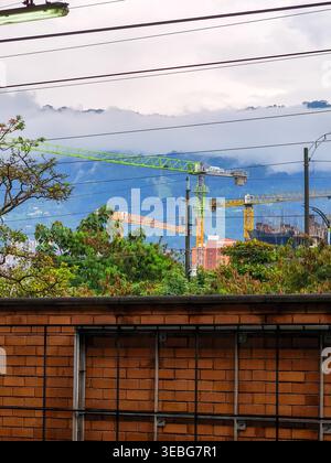 Stadtbaukrane erheben sich über Bäumen und Ziegeldächern mit nebeligen Bergen im Hintergrund und schaffen einen Kontrast zwischen Entwicklung, Natur, Stockfoto
