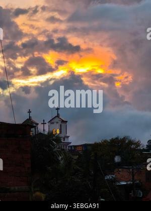 In Guatape, Antioquia, erstrahlen dramatische Sonnenuntergangswolken mit goldenem Licht über Kirchendächern und Silhouettenbäumen, die eine stimmungsvolle tropische Skyline mit sich bringen Stockfoto