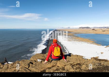 Junge Reisende, die in der Nähe einer Klippe mit Blick auf einen schwarzen Strand und den Atlantischen Ozean in Island sitzt Stockfoto