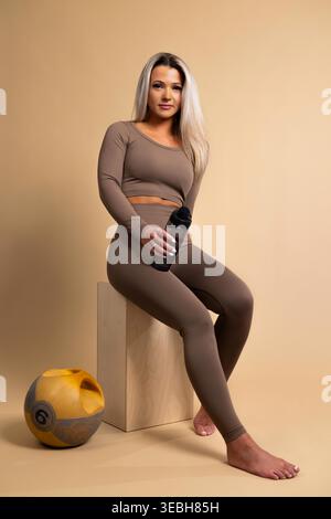 Frau in brauner Workout-Kleidung, die auf einem Holzblock sitzt und eine Wasserflasche mit einem Medizinball in der Nähe hält Stockfoto