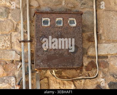 Rostige elektrische Schalttafel, die an einer Steinmauer befestigt ist, mit sichtbarer Verkabelung und verwittertem Aussehen, die industrielle und historische Saibling hervorhebt Stockfoto