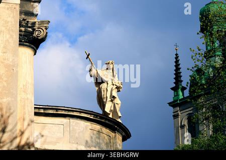 Eine verwitterte Steinstatue eines heiligen, der ein Kreuz hält, steht vor einem dramatischen Himmel, mit architektonischen Details einer Kathedrale Stockfoto
