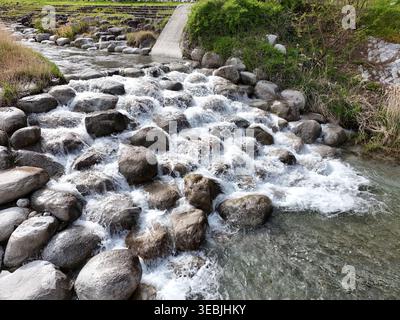 Rauschendes Wasser fließt über glatte graue Steine. Weißer Schaum tanzt, während der Strom nach unten stürzt. Grünes Gras wächst am felsigen Flussbett. Betonweg Wind Stockfoto