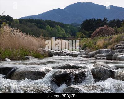 Rauschendes Wasser fließt über glatte Steine. Bäume säumen die Ufer mit herbstlichen Tönen. Der Fehler ist hinter dem fernen Hügel versteckt. Der Fluss flüstert durch Stockfoto