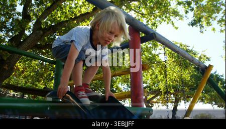 Der blonde Junge in der Hocke, der Rand der dunklen Rutsche auf dem Spielplatz unter Bäumen, blaue T-Stück rote Schuhe Stockfoto