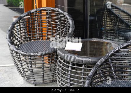 Schwarze Korbstühle und Tisch im Freien an einem Glasfenster mit Blumenarrangements im Inneren, die modernes Möbeldesign mit dekorativem Fliff kombinieren Stockfoto