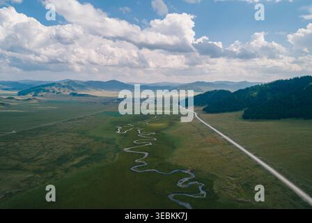 Aus der Vogelperspektive auf einen sich windenden Fluss und eine lange Straße, die durch die grünen Altai-Ebenen führt und an einem klaren Sommertag zu einem entfernten Dorf führt Stockfoto