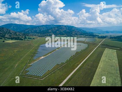 Aus der Vogelperspektive ist eine weitläufige Solarfarm-Anlage, die sich über ein grünes Tal in der Nähe majestätischer Berge erstreckt und die Sonnenenergie einfängt Stockfoto