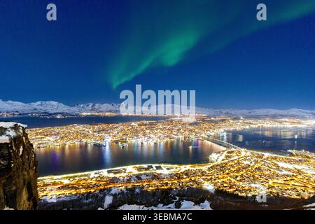 Blick auf eine atemberaubende Winternacht über einer weitläufigen Stadt, deren goldene Lichter auf dem dunklen Wasser unter den faszinierenden grünen Northern Lights, T Stockfoto