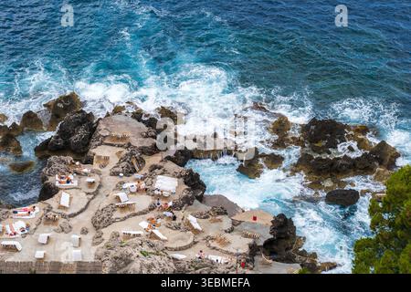 Blick auf Sonnenanbeter, die auf felsigen Terrassen liegen, während türkisfarbene Wellen gegen die zerklüftete Küste von Capri, Kampanien, Italien, krachen. Stockfoto