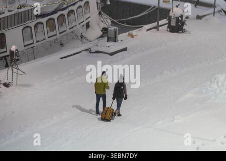 Blick auf ein Paar, das durch den frischen, dicken Schnee geht und einen gelben Koffer entlang der Straße in der Nähe eines Kanals in Tallinn, Estland, zieht. Stockfoto