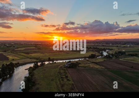 Aus der Vogelperspektive auf den farbenfrohen Sonnenuntergang über dem Morpeth River und dem umliegenden Ackerland in New South Wales, Australien. Malerische ländliche Landschaft mit lebendigem Himmel Stockfoto