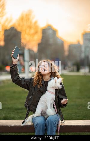 Junge Frau mit lockigem Haar filmt Video mit Telefon und kleinen Hund auf Bank Stockfoto