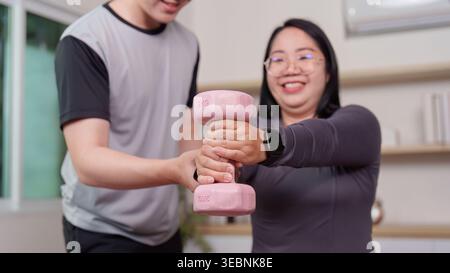 Asiatischer Mann, der einer großen Frau bei Hantelübungen zu Hause hilft Stockfoto