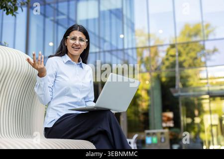 Junge indische Geschäftsfrau, die draußen auf einer Bank sitzt und sich verwirrt und gestresst fühlt, während sie auf den Laptop-Bildschirm blickt und Gesten macht, um einen zu finden Stockfoto