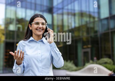 Professionelle junge Geschäftsfrau, die draußen steht, glücklich auf dem Handy spricht, wichtige Geschäfte bespricht, während sie ihre Hand in der Nähe hält Stockfoto