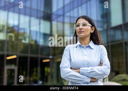 Selbstbewusste Geschäftsfrau, die mit gekreuzten Armen in die Ferne blickt, ein schickes Hemd und eine Brille trägt und sich draußen vor einem posiert Stockfoto
