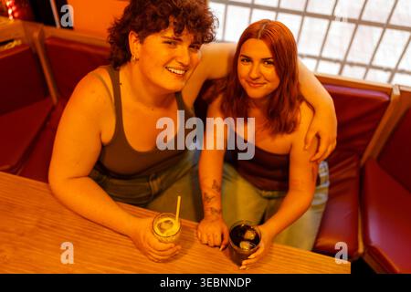 Zwei lächelnde junge Frauen an einem Bartisch, eine mit Arm um einen Freund, hält Getränke mit Eis, feiert Freundschaft Stockfoto