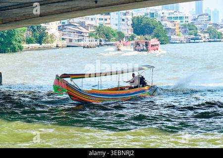 BANGKOK, THAILAND - 27. Februar 2025: Bootsfahrer an der Spitze eines dekorierten Langboot-Bootes auf dem geschäftigen Chao Phraya River mit Bangkok Skyline hinter hochwertigem ph-wert Stockfoto