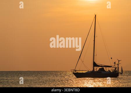 Silhouette einer Segelyacht auf ruhigem Meer bei goldenem Sonnenuntergang, friedliche abendliche Meereslandschaft mit warmer Lichtreflexion auf dem Wasser Stockfoto
