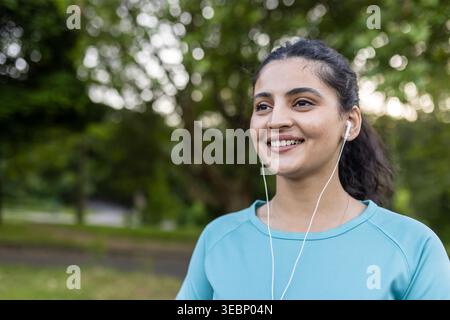 Junge indianerin lächelt fröhlich im Freien, hört Musik mit Ohrhörern, genießt Fitness und einen gesunden Lebensstil in einer grünen Park-Umgebung Stockfoto