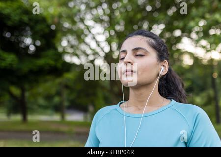 Junge indianerin mit Ohrstöpseln und einem blauen T-Shirt, die ihre Augen konzentriert schließt, friedlich in einer grünen Parkumgebung meditiert, Entspannung findet und Achtsamkeit übt Stockfoto