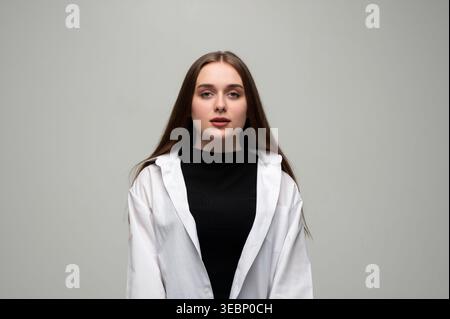 Junge Frau mit langen braunen Haaren, die selbstbewusst in weißem Hemd und schwarzem Oberteil posiert und direkt in die Kamera blickt Stockfoto