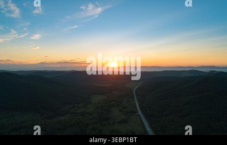 Aus der Vogelperspektive fängt ein lebhafter Sonnenuntergang über üppig grüne Berge und gewundene Straße in einem ruhigen Tal in der Abenddämmerung ein Stockfoto