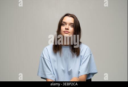 Junge Frau mit nachdenklichem Ausdruck, die ein blaues T-Shirt trägt und in einem Studio-Porträt vor grauem Hintergrund nach vorne blickt Stockfoto