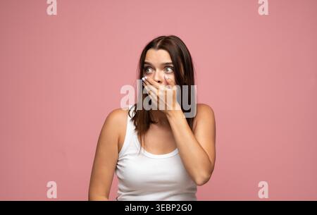 Junge Frau, die den Mund vor Schock und Überraschung mit breiten Augen bedeckt, die Emotionen auf rosa Hintergrund ausdrückt Stockfoto