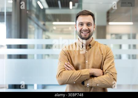 Selbstbewusster junger Geschäftsmann, der lächelnd steht, mit gekreuzten Armen steht und Professionalität und Erfolg in einer hellen, modernen Büroumgebung darstellt, die sich für geschäftliche und geschäftliche Themen eignet Stockfoto