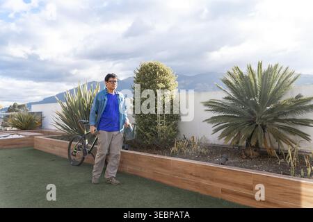 Mann steht mit Fahrrad und Wasserkanne auf einer grünen Dachterrasse mit Bergen im Hintergrund Stockfoto