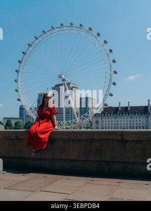 Der Blick auf eine Frau in einem leuchtend roten Kleid sitzt anmutig auf einem Steinvorsprung, hinter ihr ragt das legendäre London Eye vor einem klaren blauen Himmel, London, E Stockfoto