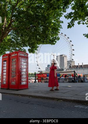 Der Blick auf leuchtend rote Telefonboxen steht im Kontrast zu den üppig grünen Bäumen, während eine Frau in einem roten Kleid mit dem London Eye im Hintergrund posiert, London, eng Stockfoto