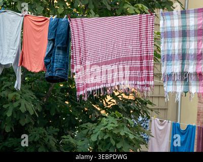 Authentische häusliche Szene mit frisch gewaschener, farbenfroher Wäsche, die im Garten an der Wäscheleine hängt, mit roten karierten Handtüchern und Jeans aus Denim Stockfoto