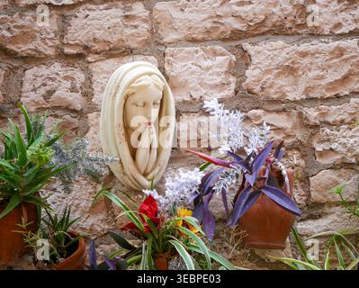 Heitere Skulptur aus weißem Marmor, betend auf einer rustikalen Steinmauer, umgeben von bunten Blumen und grünen Pflanzen in einem mediterranen Garten Stockfoto