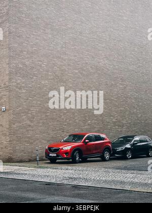 Zwei Autos parkten in der Nähe der Backsteinwand des Gebäudes. Roter Mazda-SUV und schwarzer Opel-Kombi parken vor einer hellbraunen Ziegelmauer. Stockfoto