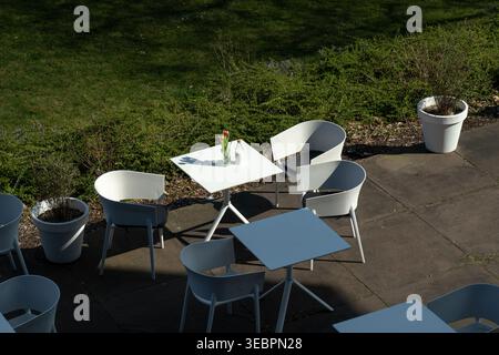 Blick von oben auf einen leeren Sitzbereich im Freien mit weißen quadratischen Tischen und weißen Stühlen auf einer Steinterrasse neben einem grasbewachsenen Bereich mit Topfpflanzen Stockfoto